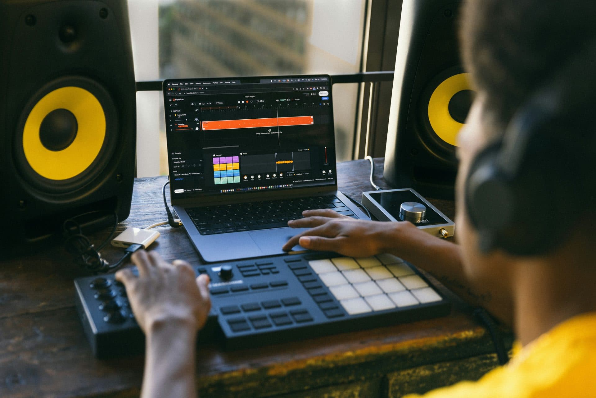 A producer working on a laptop and pad controller with KRK Rokit monitors, headphones on, a DAW arrangement view on screen